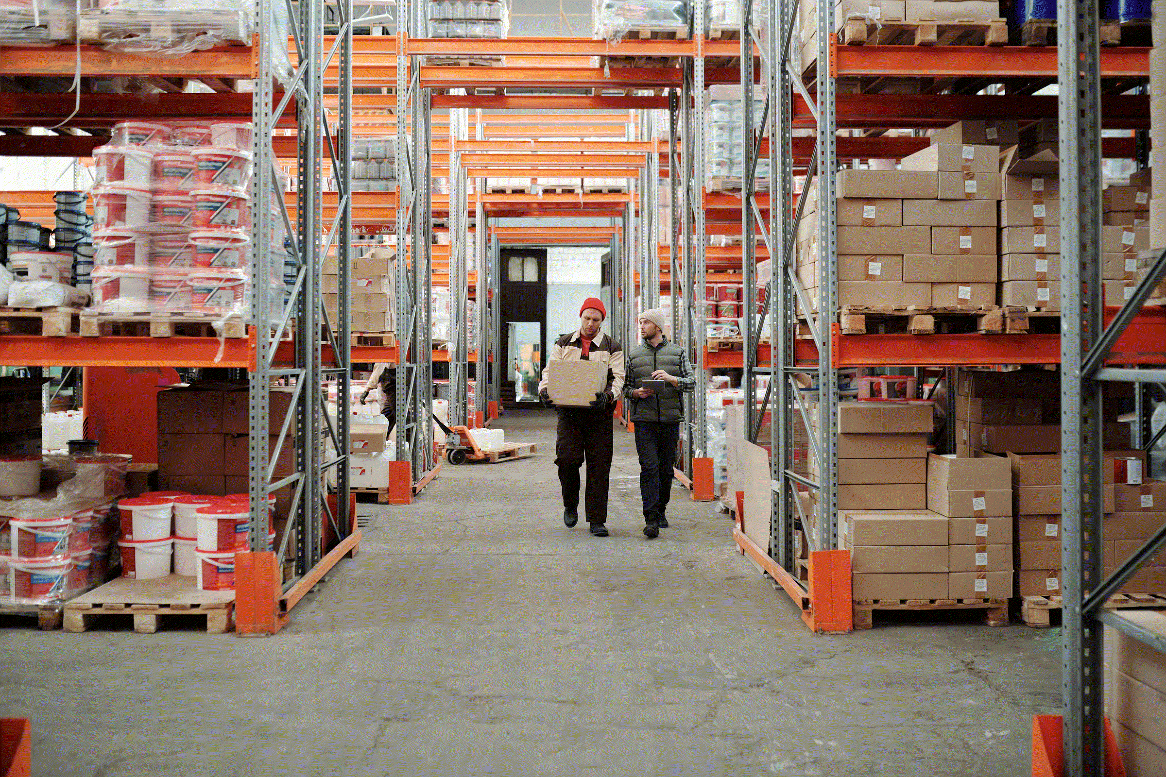 Two warehouse workers walking between tall pallet racks, one carrying a box